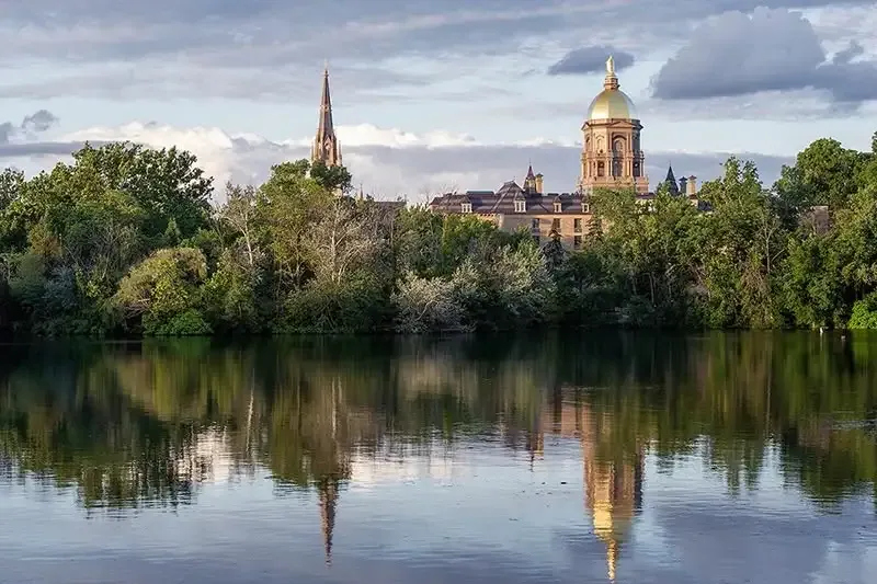 The University of Notre Dame's Golden Dome and Basilica of the Sacred Heart are reflected in St. Joseph's Lake. Lush green trees line the lake's edge. A partly cloudy sky adds depth to the serene scene.